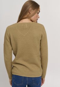 Knit sweater in light brown, featuring a textured pattern, long sleeves, and a wide neckline, paired with blue jeans.