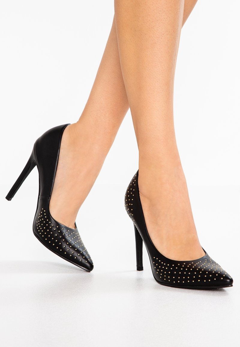 Jambes portant des talons hauts noirs avec des clous dorés, des bouts pointus et des talons aiguilles, posées sur une surface blanche.