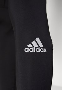 Czarne legginsy sportowe o gładkiej fakturze z wyraźnym białym logo Adidas na lewej udzie. Dopasowany krój.