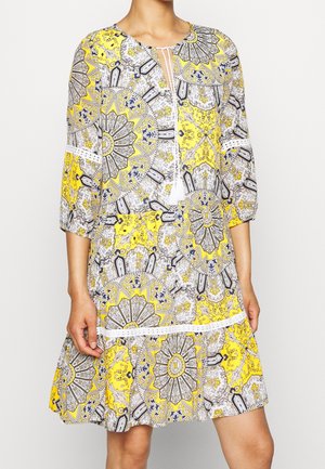 Robe à motifs jaunes, blancs et noirs jusqu'aux genoux avec manches 3/4, col rond et détail noué à l'avant portée par une personne.