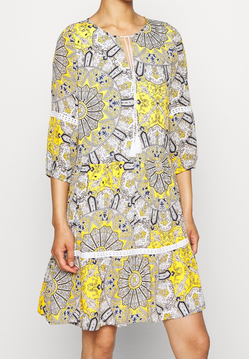 Robe à motifs jaunes, blancs et noirs jusqu'aux genoux avec manches 3/4, col rond et détail noué à l'avant portée par une personne.