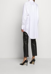 Persona vestida con una camisa larga blanca y holgada, pantalones de cuero negros cortos y zapatos de tacón alto negros, de pie contra un fondo liso.