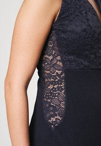 Marindress med en figursydd design och spetsdetaljer på sidorna, som visar blommönster och en mjuk tygtextur.