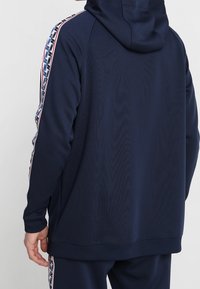 Marininblå zip-up hoodie med texturerad yta, med kontrasterande randigt mönster längs ärmarna och en känguruficka.