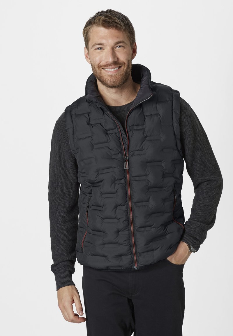 Schwarze Steppweste mit einem gesteppten Design, Reißverschluss vorne und Seitentaschen. Getragen über einem dunklen Pullover mit hohem Kragen und strukturierten Ärmeln.