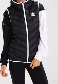 Černobílá péřová vesta se vzorem ve tvaru šipky, s kapucí, předním zipem a třemi bílými pruhy na rukávech. Na hrudi logo Adidas.