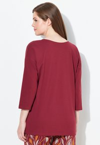 Vrouw met lang bruin haar draagt een losse, donkerrode top met driekwartmouwen en een patroon van oranje, wit en kastanjebruine broek, staand tegen een witte muur.