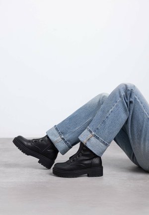 Stivaletti neri in pelle con finitura texturizzata e punta tonda. Caratterizzati da una suola robusta e un design con lacci, abbinati a jeans azzurri scampanati.