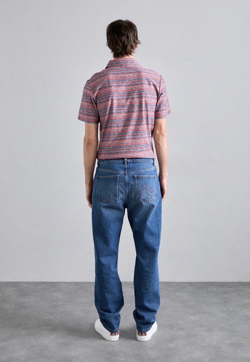 Randig polo skjorta i röd, blå och vit; klassiska blå denimjeans; vita sneakers med mönstrade accenter; sedd bakifrån.