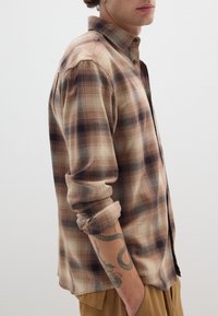 Camicia a righe marrone e crema con bottoni, realizzata in tessuto morbido, con maniche arrotolate e due polsini con bottone; presenta un colletto e un orlo laterale.