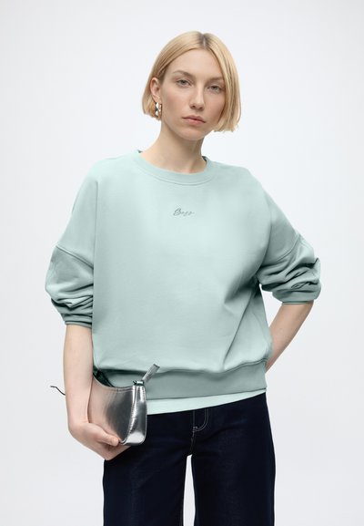 Femme aux cheveux blonds courts portant un sweat-shirt bleu clair et un pantalon foncé, tenant une petite pochette argentée.