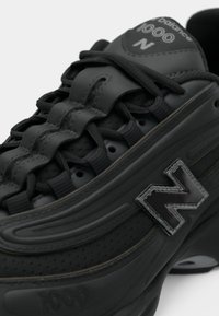 Chaussure de sport noire avec un matériau texturé, présentant un design incurvé, des lacets proéminents et un logo "N" brillant sur le côté.
