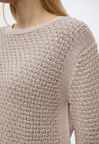 Κοντινό πλάνο μπεζ πλεκτού πουλόβερ με περίπλοκη υφή ραφής στον ώμο, το στήθος και το μανίκι, που φοριέται από άτομο με ξανθά μαλλιά.