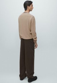 Beige stickad tröja med rund halsringning, kombinerad med löst sittande bruna byxor. Texturerat tyg och enkel design. Svarta skor syns.