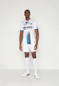 Άνδρας ντυμένος με λευκή στολή ποδοσφαίρου με μπλε και μαύρες κάθετες ρίγες, λευκές κάλτσες και παπούτσια με καρφιά μπροστά σε απλό φόντο.