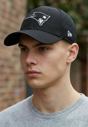 Junger Mann mit schwarzer New England Patriots-Cap und grauem Hemd, steht im Freien vor einem unscharfen Hintergrund aus einer Backsteinwand.