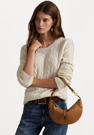 Barna bőr kézitáska, ívelt formával és aranyszínű fémrészletekkel, krém színű kötött pulóver és kék farmernadrág fölött hordva.