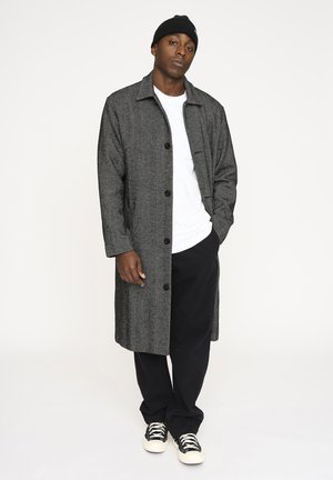 Mand iført en grå lang frakke over en hvid skjorte, sorte bukser, sort hue og sorte og hvide sneakers, stående med den ene hånd i lommen.
