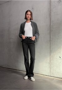 Jeune femme se tient contre un mur en béton, portant une veste grise, une chemise blanche, un jean noir et des baskets blanches, les mains dans les poches.