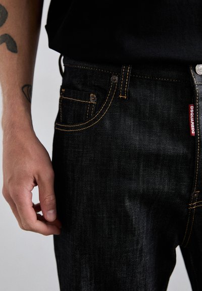 Main gauche près de la hanche d'une personne portant un jean Dsquared2 noir avec des coutures oranges visibles et des tatouages sur l'avant-bras.