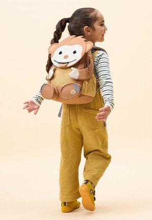 Child facing sideways wearing yellow overalls, striped long-sleeve shirt, and a playful monkey-shaped backpack, walking with arms outstretched.
