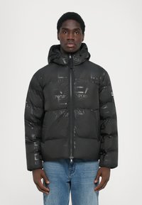 Schwarze Steppjacke mit glänzender Oberfläche, ausgestattet mit einem Reißverschluss vorne, Kapuze und geprägten Logo-Mustern. Strukturierte, isolierte Ausführung.