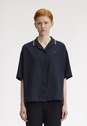 Jeune personne aux cheveux roux courts portant une chemise ample à manches courtes bleu marine foncé avec col et détail rayé, debout devant un fond blanc uni.