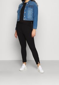Veste en denim avec fermeture à boutons, deux poches poitrine et manches retroussées. Leggings noirs avec fentes sur les côtés et baskets blanches.