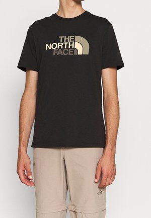 Mand iført sort The North Face t-shirt og beige bukser med The North Face-logo nær lommen, stående mod en ensfarvet baggrund.