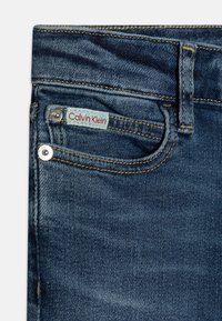 Jeans in denim blu scuro con una tasca frontale che presenta un'etichetta Calvin Klein rossa e azzurro chiaro ben visibile, bottone argento e cuciture gialle.