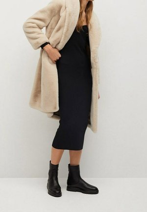 Femme portant un manteau beige duveteux, une robe midi noire sans manches et des bottines noires, debout contre un mur blanc uni.