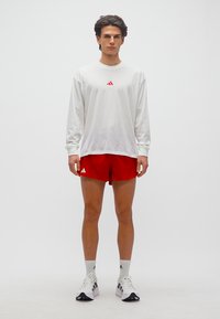 Νέος άνδρας όρθιος, φορώντας λευκό πουκάμισο με μακριά μανίκια, κόκκινα σορτς, λευκές κάλτσες και λευκά αθλητικά παπούτσια σε απλό λευκό φόντο.