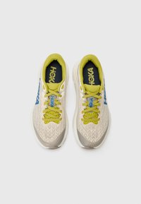 Könnyű sneaker, bézs színű hálós felsőrésszel, sárga cipőfűzőkkel, kék logóval és vastag fehér talppal, a kényelem és a támogatás érdekében tervezve.