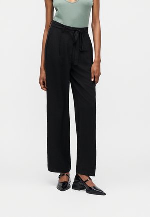 Žena oblečená v čiernych širokých nohaviciach s vysokým pásom a látkovým opaskom, zelený bezrukávový top a čierne špicaté topánky Mary Jane s masívnym podpätkom.