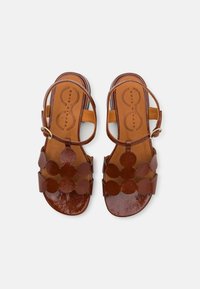 Sandales en cuir marron avec une finition lisse, présentant des découpes circulaires sur les lanières et une fermeture à boucle. Design à semelle plate.