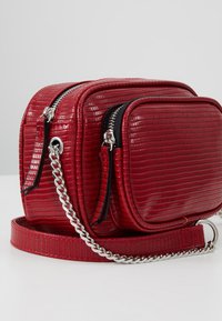 Borsa a tracolla rossa in pelle sintetica con superficie testurizzata, due scomparti con zip e una tracolla in catena argentata.