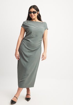 Long, fitted dress in textured olive green fabric, featuring short sleeves and a draped design. Paired with black pointed flats and sunglasses.
