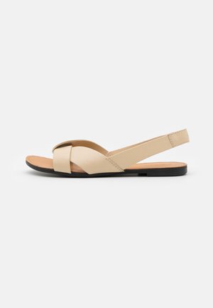 Beige flad sandal med krydsede frontstropper og slingback-strop, tan indersål og sort ydersål, vist fra siden på hvid baggrund.