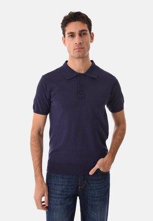 Uomo con capelli corti e scuri che indossa una polo a maniche corte blu navy e jeans blu scuro, in piedi davanti a uno sfondo chiaro e uniforme.