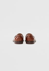 Paire de mocassins en cuir marron poli, vues de dos sur un fond gris clair uni.