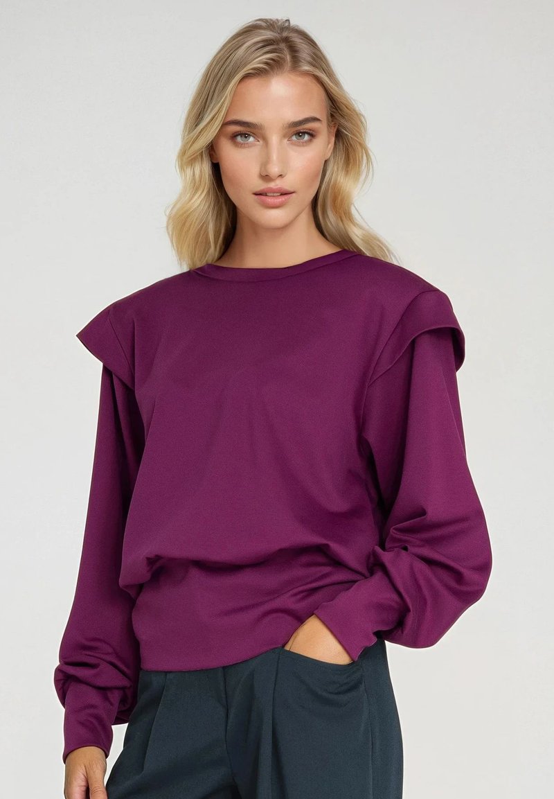 Fioletowy długi rękaw bluza z falbanami na ramionach, okrągłym dekoltem i miękką teksturą. Połączona z ciemnozielonymi spodniami.