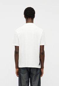 Άνδρας με σκούρο δέρμα και κοντά, σγουρά μαλλιά, στέκεται με την πλάτη προς τα εμπρός, φορώντας ένα απλό λευκό κοντομάνικο μπλουζάκι και σκούρο τζιν, μπροστά σε λευκό φόντο.