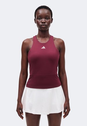 Femme portant un débardeur de sport bordeaux avec un logo Adidas blanc et une jupe de sport plissée blanche, debout devant un fond uni.