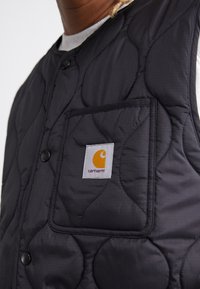 Black quilted vest with a textured pattern, featuring a chest pocket and a rectangular logo patch in orange and white on the left side.