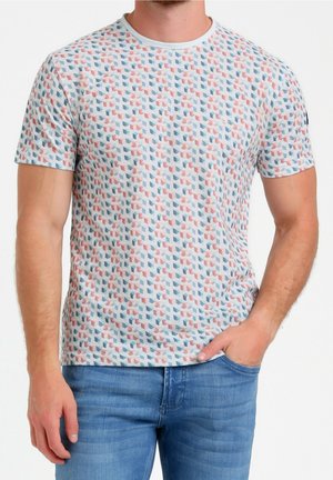 Mężczyzna ubrany w biały T-shirt z krótkim rękawem, z czerwono-niebieskim geometrycznym wzorem, w niebieskich jeansach, z prawą ręką w kieszeni, stojący na białym tle.