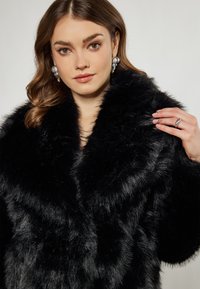 Czarny płaszcz z sztucznego futra o szerokim kołnierzu i miękkim, teksturowanym wykończeniu. Ręka modelki spoczywa na kołnierzu, ukazując szczegółowy pierścionek.