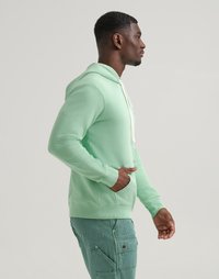 Uomo che indossa una felpa di colore verde menta con lacci bianchi e pantaloni cargo turchesi, in piedi di lato con una mano nella tasca della felpa.