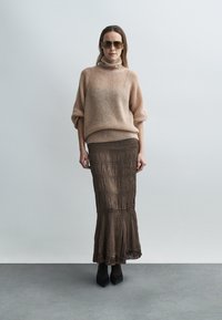 Pull en maille beige à col montant, associé à une jupe plissée marron mi-mollet. Le modèle porte des lunettes de soleil foncées et des talons noirs.