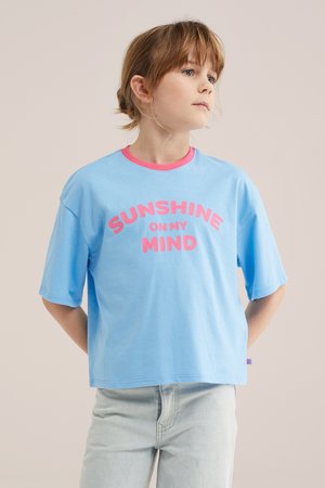 Nooruk, kellel on valgelt pruuni värvi juuksed seotud tagasi, kannab sinist T-särki roosade sõnadega "SUNSHINE ON MY MIND" ja heledeni teksapükse, vaadates küljele.