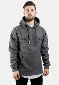 Sudadera con capucha gris que cuenta con un bolsillo frontal y un cordón de ajuste, con un logo mate en el centro. La tela parece tener una textura suave.